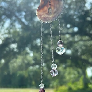 May include: A seashell car charm with dangling chains and crystal beads. The shell is a light brown color, and the chains are silver. The beads are clear and purple. The charm hangs in front of a blurred green background.