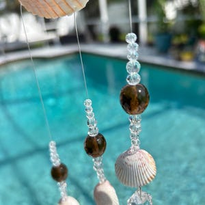 May include: A seashell wind chime with clear beads and brown, speckled beads. The chime features various seashells and is suspended by clear string. The background shows a swimming pool.