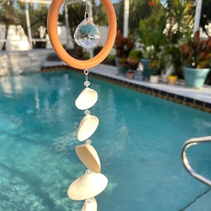 May include: A seashell wind chime, featuring a wooden ring and a crystal ball, hangs in front of a swimming pool. The chime comprises several white seashells strung together with clear beads. The wooden ring is suspended from a chain. The background shows greenery.