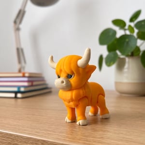 May include: A small, orange figurine of a Highland cow with white horns and hooves. The toy cow is standing on a wooden surface. The figurine has a detailed design and is a cute decorative item.