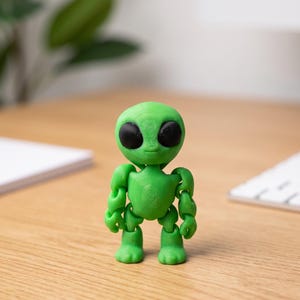 Op de afbeelding: Een groene, beweegbare alien figuur met grote zwarte ogen. De speelgoed alien heeft een ronde kop, een klein lichaam en beweegbare armen en benen. Het beeldje staat op een houten oppervlak.