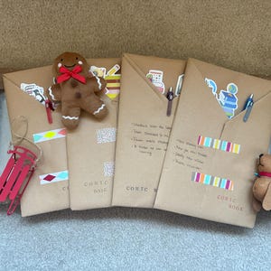 May include: Four brown paper comic book covers decorated with stickers, pens, and colorful tape. A gingerbread man, a red sled, and a stuffed dog are placed on the covers. The words "COMIC BOOK" are printed on the covers.