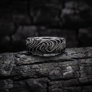 Könnte beinhalten: Ein dunkel silberfarbener Ring mit einer tief texturierten, wirbelnden, wellenartigen Struktur auf der gesamten Außenfläche. Das Metall hat ein mattes, verwittertes Finish mit kontrastreichen Schatten, die die eingravierten Rillen betonen.