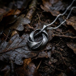 May include: A silver-toned metal pendant featuring an intricate, twisted knot design with a textured, matte finish. The pendant is attached to a delicate, matching silver-toned metal chain that extends upward from the top of the knot structure.