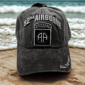 May include: A charcoal gray baseball cap with the text "82nd AIRBORNE" embroidered in white. The cap features a patch with "AIRBORNE" above a black square with "AP" inside. The brim has "ALL THE WAY!" embroidered in white.