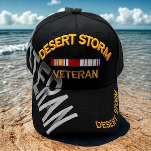 Könnte beinhalten: Schwarze Baseballkappe mit gold- und weißgesticktem Text "DESERT STORM VETERAN". Mit einem Banddesign und dem Wort "VETERAN". Die Kappe befindet sich auf einem sandigen Strand mit blauem Ozean im Hintergrund.