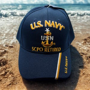 Könnte beinhalten: Marineblaue Baseballkappe mit gold- und weißgesticktem Text und Grafiken. Der Text lautet "U.S. NAVY", "USN" und "SCPO RETIRED". Ein gold-weißer Streifen verläuft die Seite der Kappe hinunter, mit "U.S. NAVY" auf der Seite gestickt.