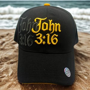May include: Black baseball cap with the text "John 3:16" embroidered in yellow and gray. The cap has a yellow brim and button. A small, circular sticker is on the side.
