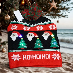 May include: A festive Christmas beanie in black, red, and white, with a green and red pom-pom. The hat displays Santa faces, Christmas trees, and the text "HO! HO! HO!". The hat has small lights and is on a sandy beach.