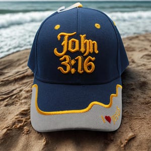 May include: A navy blue baseball cap with the text "John 3:16" embroidered in gold. The cap has a gray brim with yellow trim and a small red heart with the word "Jesus" embroidered on the side. The cap is sitting on sand.