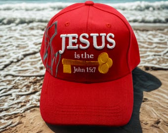 May include: A red baseball cap with the text "JESUS is the John 15:7" embroidered in white and gold. The cap features a curved brim and a six-panel design. The background shows a sandy beach and ocean waves.