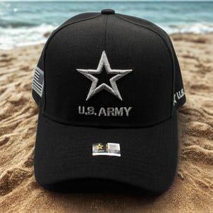 Op de afbeelding: Zwarte baseballcap met een wit geborduurde ster en de tekst "U.S. ARMY". De cap heeft een Amerikaanse vlag patch aan de zijkant en een label op de rand. De cap ligt op een zandstrand.