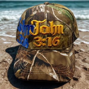 以下が含まれることがあります： ゴールドとブルーの糸で「John 3:16」と刺繍された迷彩柄の野球帽。帽子はクラシックな6パネルのデザインとカーブしたつばが特徴です。背景には砂浜と海が写っています。