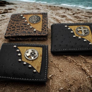 May include: Three bi-fold wallets featuring textured leather panels in black and brown. Each wallet has a tan inlay, a row of silver-toned metal studs along the seam, and a circular silver-toned metal emblem depicting a cross, a star, or a figure.