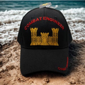 May include: Black baseball cap with "COMBAT ENGINEER" embroidered in red, and a gold castle emblem. The cap is on a sandy beach with the ocean in the background. A tag is attached to the cap.