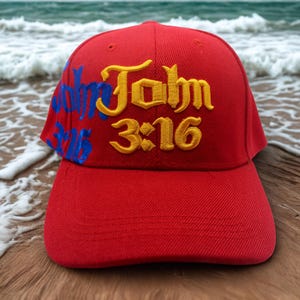 May include: A red baseball cap with the text "John 3:16" embroidered in yellow and blue. The hat is set against a backdrop of a beach with ocean waves and wooden planks.