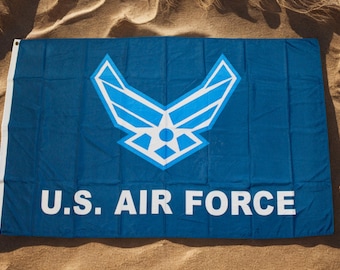 Puede incluir: Una bandera azul con las palabras "U.S. AIR FORCE" en letras blancas. La bandera presenta el símbolo de la Fuerza Aérea de EE. UU. en blanco y azul claro. La bandera rectangular está sobre una superficie arenosa.