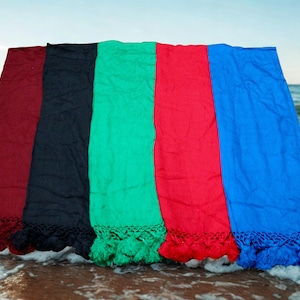 Op de afbeelding: Vijf kleurrijke stranddoeken in de kleuren bordeaux, zwart, groen, rood en blauw, met gefranjerde randen. De doeken worden gepresenteerd op een zandstrand met oceaangolven op de achtergrond.