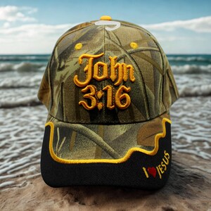 Peut inclure: Casquette de baseball camouflage avec le texte "John 3:16" brodé en fil doré. La visière noire porte l'inscription "I ❤️ Jesus" en or et un cœur rouge. La casquette est posée sur une plage.