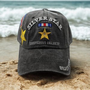 May include: A gray baseball cap with the words "SILVER STAR" embroidered on the front. The cap features a gold star and the text "CONSPICUOUS GALLANTRY". A red, white, and blue ribbon is also embroidered on the cap.