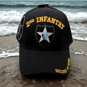 Könnte beinhalten: Schwarze Baseballkappe mit gesticktem Text und Wappen. Die Kappe trägt die gelben Worte "2ND INFANTRY", ein weißes Sternemblem und den gelben Schriftzug "SECOND TO NONE". Die Kappe ist vor einem verschwommenen Ozeanhintergrund platziert.