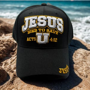 Puede incluir: Gorra de béisbol negra con el texto "JESUS DIED TO SAVE" bordado en blanco y dorado. La gorra también presenta "ACTS U 4:12" y "JESUS" bordados en el lateral. La gorra está sobre un fondo de playa.