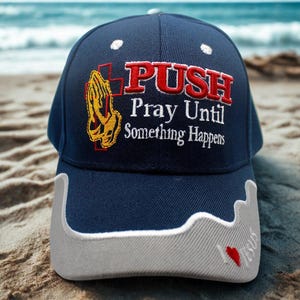 May include: Navy blue baseball cap with the text "PUSH Pray Until Something Happens" in red and white. The cap features an embroidered praying hands design in yellow and a gray bill with a red heart.