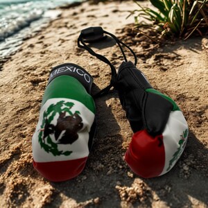 May include: A pair of miniature boxing gloves featuring the green, white, and red colours of the Mexican flag, with the word MEXICO printed in white on the black wrist cuff and a detailed eagle emblem on the white section of the gloves.