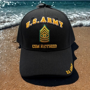 Puede incluir: Gorra de béisbol negra con texto dorado bordado y una insignia militar. El texto en la gorra dice "U.S. ARMY" y "CSM RETIRED". La gorra está sobre una playa de arena, con el mar al fondo.
