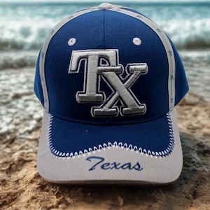 May include: A navy blue baseball cap with a grey brim and accents. The cap features the letters "TX" in a bold, outlined design. The word "Texas" is embroidered on the brim. The cap is set against a beach background.