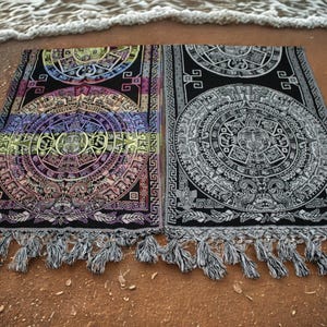 Op de afbeelding: Twee rechthoekige strandlakens met Azteekse kalenderontwerpen. Eén handdoek heeft een kleurrijk ontwerp met paarse, gele en blauwe tinten, terwijl de andere zwart-wit is. Beide hebben franjes en liggen op een zandstrand.