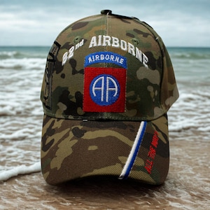 May include: Camouflage baseball cap with the text "82nd AIRBORNE" in white lettering. The cap features a patch with "AIRBORNE" and "AP" in a blue and red design. The brim has the text "ALL THE WAY" in red.