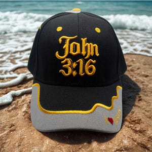May include: Black baseball cap with the text "John 3:16" embroidered in gold. The cap has a gray brim with a gold outline and a small red heart. The background shows a sandy beach and ocean waves.