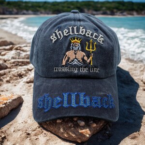 Peut inclure: Casquette de baseball bleu marine avec le mot "shellback" brodé en blanc au-dessus d'une illustration d'une figure musclée avec un trident. L'expression "crossing the line" est en dessous, et "shellback" est brodé en bleu sur la visière.