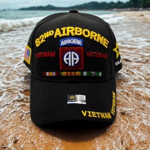 May include: Black baseball cap with embroidered text and patches. The cap features "82nd Airborne", "Vietnam Veteran", and a military emblem. A small American flag patch is on the side. The cap is on a sandy beach.