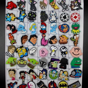 May include: A collection of colorful shoe charms featuring various cartoon characters, logos, and designs. Includes characters like Tom and Jerry, Snoopy, Batman, and the Smurfs. These charms are designed to be attached to shoes.