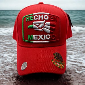 May include: A red baseball cap with a curved peak. The cap displays a patch with the words "HECHO EN MEXICO" and a Mexican flag design. A smaller emblem of the Mexican coat of arms is on the side. The cap is set against a beach backdrop.
