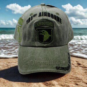 Puede incluir: Gorra de béisbol verde oliva con el texto "101st Airborne" y un parche con un águila. La gorra tiene una visera curva y las palabras "Screaming Eagles" están bordadas en el lateral. Fondo: playa y mar.