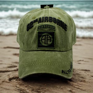 May include: Olive green baseball cap with the text "82nd AIRBORNE" in black, along with a patch featuring "AIRBORNE" and "AP" in a circle. The phrase "ALL THE WAY?" is embroidered on the brim. The cap is on a sandy surface.