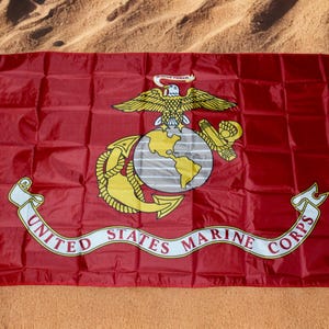 Op de afbeelding: Een rode vlag met het embleem van het United States Marine Corps. Het embleem toont een adelaar, wereldbol en anker, met de tekst "United States Marine Corps" op een witte banner. De vlag ligt op zand.