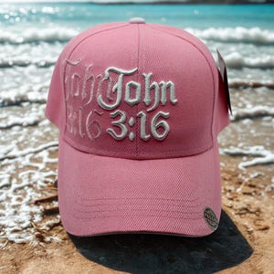 May include: A pink baseball cap with white embroidered text that reads "John 3:16". The cap has a curved brim and a button on top. The background shows a beach and ocean.