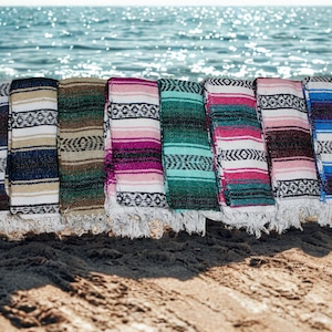 Może przedstawiać: Kolekcja kolorowych, tkanych koców z frędzlami, wystawionych na piaszczystej plaży z oceanem w tle. Koce mają różne wzory w paski w odcieniach niebieskiego, zielonego, różowego i brązowego.