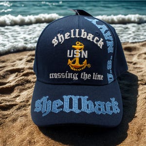 Peut inclure: Casquette de baseball bleu marine avec le mot "shellback" brodé en blanc et bleu clair. La casquette présente le logo USN avec une ancre dorée et la phrase "crossing the line". La casquette est sur une plage de sable.