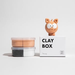 May include: A clay box set with a pig figurine. The box is white with the words "CLAY BOX" and "The pig" printed on it. Two small plastic containers of clay are stacked below the box. The pig figurine is light brown with large white eyes.