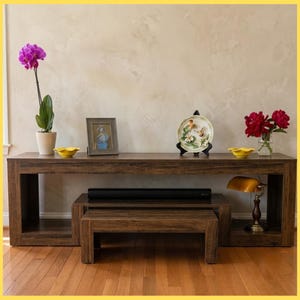 May include: A dark wood console table with a matching low bench underneath. The table holds a purple orchid in a white pot, a framed picture, a decorative plate with bird imagery, two yellow bowls, and a brass desk lamp with a yellow glass shade.