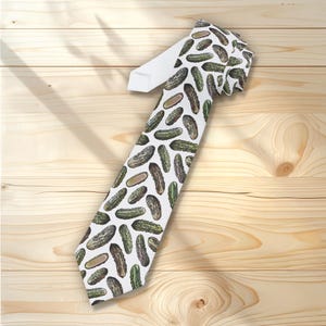 Può includere: Una cravatta bianca con un motivo di sottaceti verdi e marroni. La cravatta è realizzata in un tessuto liscio e viene esposta su una superficie di legno chiaro. Il motivo dei sottaceti si ripete su tutta la superficie della cravatta.