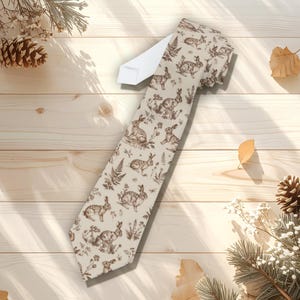 May include: A beige necktie featuring a repeating pattern of brown rabbits and foliage. The tie has a white section at the top and is displayed on a light wood surface. The design is reminiscent of a vintage illustration.