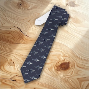 Può includere: Cravatta blu navy con un motivo ripetuto di squali bianchi e grigi. La cravatta ha un retro bianco ed è esposta su una superficie di legno. Questo accessorio è adatto per occasioni formali o informali.
