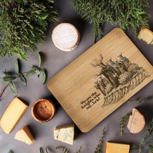 Peut inclure: Planche à découper en bambou avec une illustration humoristique et du texte, entourée de différents fromages et de verdure. La planche présente un dessin de personnages et la phrase "We are the knights who say... brie!"
