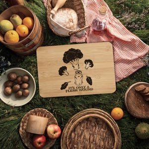 Pode incluir: Uma tábua de corte de bambu com um desenho gravado a laser de brócolis e a frase "IT'S ONLY A FLESH WOUND". A imagem também inclui elementos de piquenique, como frutas e pão.